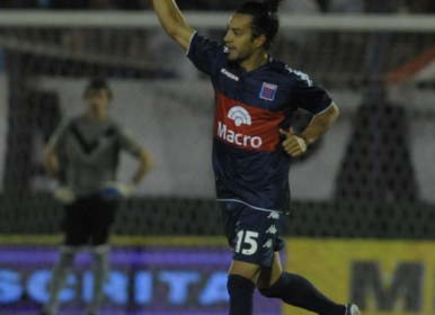 Soccer player celebrating on the ground after scoring a goal, with a blurred crowd and another person standing in the background.