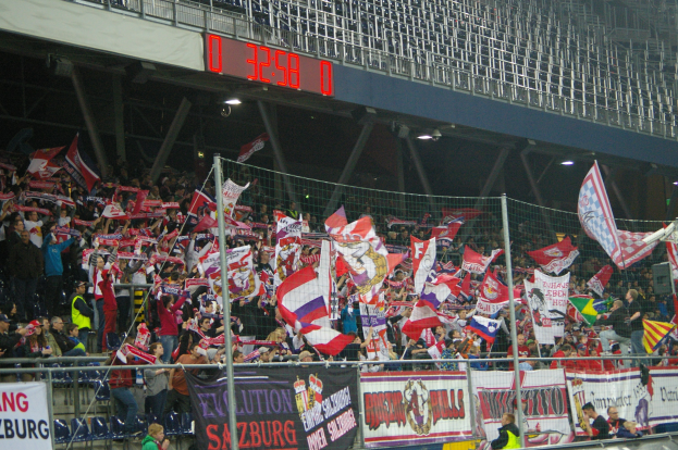 Eine große Menschenmenge in einem hell erleuchteten Stadion schwenkt Fahnen und Banner mit Text, getrennt durch ein Netz von einem digitalen Anzeigebrett oben.