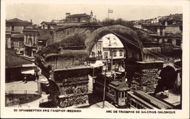 Schwarz-weiß-Foto einer Stadtstraße mit einer Bogenbrücke in der Mitte, umgeben von Gebäuden, Bäumen, Pfählen, Drähten und Menschen; Text unten.
