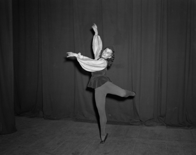 Eine Frau in einem Schwarz-Weiß-Foto steht auf einer Bühne mit einem Vorhang im Hintergrund, trägt Schuhe und ist bereit zur Vorstellung.