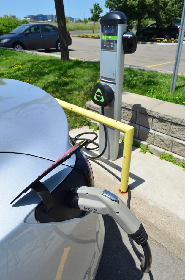 Elektroauto-Laden an einer Straßenstation mit Gras, Bäumen, Strommasten, Gebäuden und einem klaren blauen Himmel im Hintergrund.