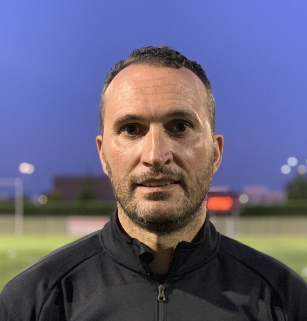 Ein Mann in einer schwarzen Jacke mit einem Logo steht auf einem Fußballfeld und sieht aus wie ein Trainer mit einem unscharfen Hintergrund.