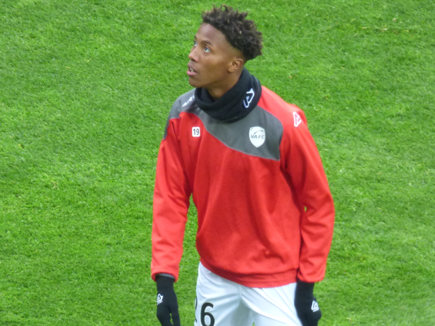 Ein junger Mann in einer roten Jacke und weißen Shorts steht auf einem Fussballfeld und schaut rechts mit entschlossener Miene, dabei Baseball-Handschuhe tragend.