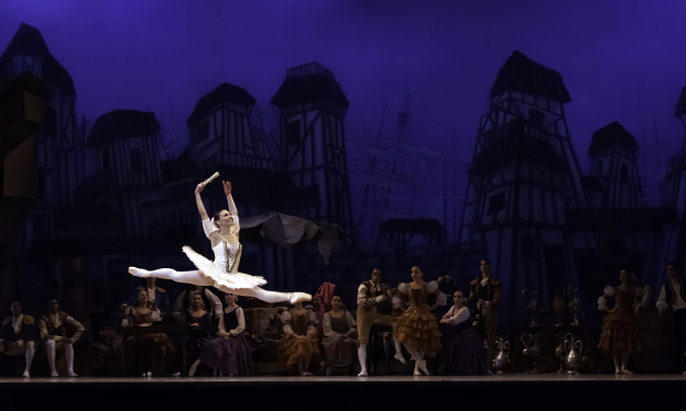 Eine Balletttänzerin in einem weißen Tutu und Spitzenschuhen tanzt auf der Bühne, während das Publikum zusieht, mit einem Burgenbild im Hintergrund.