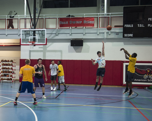 Eine Gruppe von Männern, die Basketball in einer Turnhalle mit einem Korb, Netz, Wand und Anzeigetafel spielen.