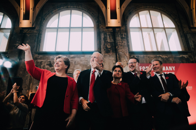 Eine Gruppe von Menschen, wahrscheinlich Labour-Parteimitglieder, die jubelnd vor einer Menge stehen, mit einem Banner und beleuchteten Glasfenstern im Hintergrund.