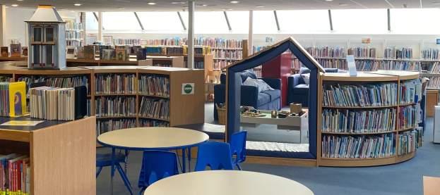 Eine geräumige Bibliothek mit Bücherregalen, Tischen, Stühlen, Sofas mit Kissen, Deckenleuchten und einem zentralen Pfosten.