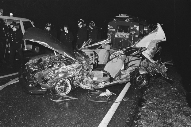 Schwarz-weiß-Foto eines beschädigten Autos am Straßenrand mit mehreren Personen, einige mit Helmen, in der Nähe und einem Fahrzeug im Hintergrund.