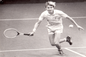 Ein junger Mann in weißer Tenniskleidung schlägt einen Ball auf einem Tennisplatz mit einem entschlossenen Gesichtsausdruck.