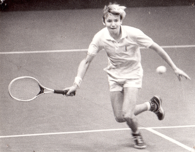 Ein junger Mann in weißer Tenniskleidung schlägt einen Ball auf einem Tennisplatz mit einem entschlossenen Gesichtsausdruck.