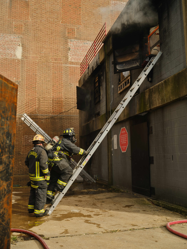 Feuerwehrleute in Helmen klettern eine Leiter zu einem rauchgefüllten Gebäude hoch, während ein metallisches Objekt und Bodenrohre sichtbar sind.