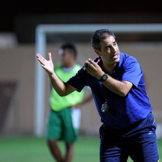 Ein Mann in einem blauen T-Shirt und Hose steht auf einem Fußballfeld und hält ein Mikrofon in der Hand, während er gestikuliert, als würde er trainieren. Im Hintergrund sind eine Person in einem grünen Kleid, ein Tor und eine Wand zu sehen.