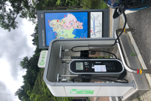 Eine Ladestation für Elektrofahrzeuge am Straßenrand mit einem Motorrad in der Nähe, ein digitales Anzeigetafel, die anzeigt, dass es eine E-Bike-Ladestation ist, mit Bäumen und einem klaren blauen Himmel im Hintergrund.