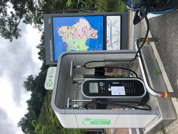 Eine Ladestation für Elektrofahrzeuge am Straßenrand mit einem Motorrad in der Nähe, ein digitales Anzeigetafel, die anzeigt, dass es eine E-Bike-Ladestation ist, mit Bäumen und einem klaren blauen Himmel im Hintergrund.