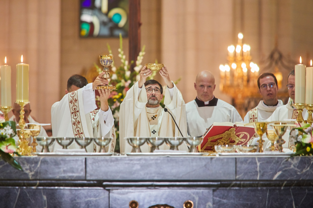 Eine Gruppe von Priestern vor einem Altar, jeder hält einen Kelch in der Hand, mit Gläsern, einem Mikrofon, einem Buch und anderen Gegenständen auf einem Tisch in der Mitte, einem Blumenstrauß links, einem Kerzenständer mit Kerzen rechts und einem Buntglasfenster im Hintergrund.