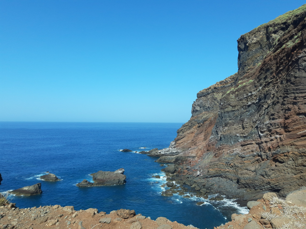 Luftaufnahme des Ozeans von einer Klippe in Lanzarote aus, mit Wasser im Vordergrund, Himmel im Hintergrund, einem Hügel auf der rechten Seite und Felsen und Gras unten.