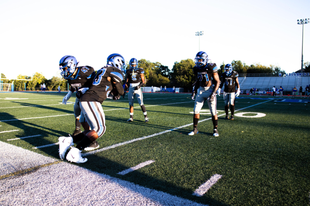 Eine Gruppe von Menschen, die Fußball auf einem Feld spielen, das von Bäumen, Laternenmasten und einem klaren blauen Himmel umgeben ist, wobei einige Spieler Helme und Schuhe tragen.