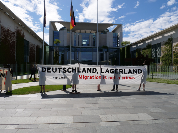 Eine Gruppe von Menschen hält ein Transparent mit der Aufschrift "Deutschland, Lagerland Migration ist kein Verbrechen" vor einem Gebäude mit Fenstern, umgeben von Gras, einem Metallzaun, Pflanzen, Bäumen und Fahnen mit Stangen unter einem bewölkten Himmel.