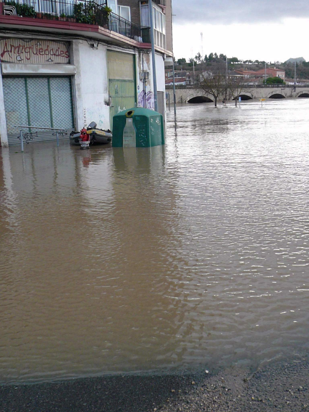 Überschwemmte Straße mit einem von Wasser umgebenen Gebäude, das ein Nachbargebäude mit Fenstern, Geländern, Pflanzen und einem Namensschild sowie im Hintergrund Bäume, eine Brücke, Pfähle, andere Gebäude und einen bewölkten Himmel zeigt.