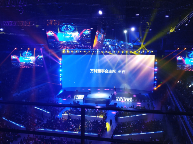 Eine große Menschenmenge sitzt vor einer hell erleuchteten Bühne mit Metallabsperrungen und wartet auf ein E-Sport-Event mit einem großen Bildschirm im Hintergrund.