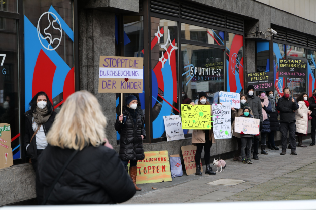 Gruppe von maskierten Menschen mit Schildern und Plakaten vor einem Gebäude mit Glastüren, einige mit Kameras und einem Hund in der Nähe, protestieren gegen ein deutsches Regierungsverbot von Masken in Schulen.