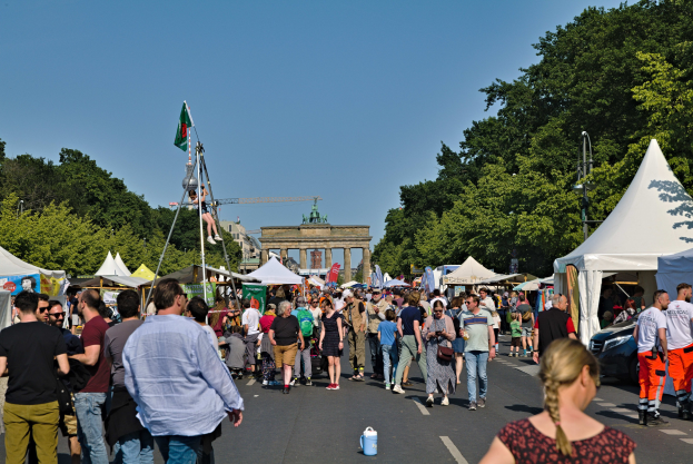 Eine Menschenmenge, die eine Straße entlanggeht, die mit Zelten, Fahrzeugen und Bäumen gesäumt ist, mit einem Bogen und einem klaren blauen Himmel im Hintergrund und Fahnenmasten auf der linken Seite.