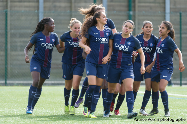 Gruppe junger Frauen, die Fußball auf einem Rasenfeld mit Maschendrahtzaun und einer Wand im Hintergrund spielen, mit dem Text "Paris Saint-Germain Women's Soccer" in der rechten unteren Ecke.