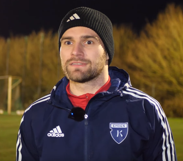 Ein Mann in einer blauen Jacke und Mütze steht auf einem Fußballfeld während eines Trainings, mit Bäumen und einem Torpfosten im Hintergrund.