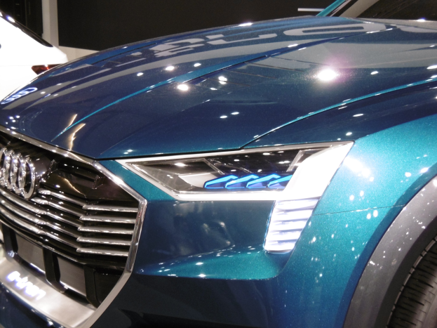 Blauer Audi e-tron Conceptwagen auf einer Automesse zu sehen, mit einem weißen Auto im Hintergrund und einer schwarzen Wand dahinter.