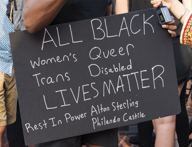 Eine Gruppe von Menschen, die zusammenstehen, wobei eine Person in der Mitte ein Schild hält, auf dem "All Black Women's Queer Trans-Disabled Lives Matter" steht, und eine andere Person ein Objekt hält, sowie Stühle im Hintergrund zu sehen sind.