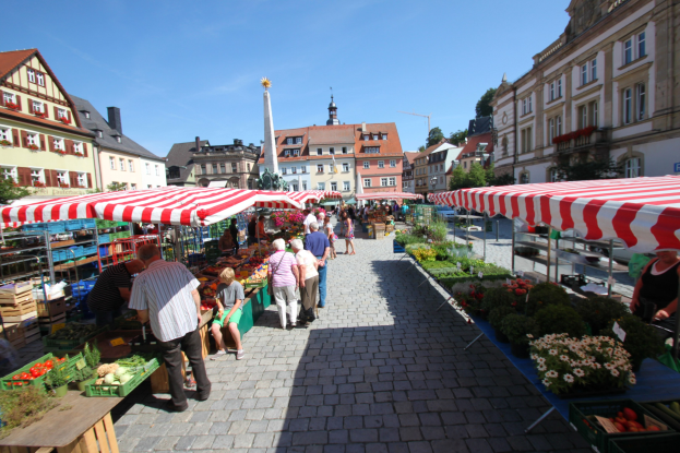 Ein belebter Markt im alten Stadtzentrum von Heidelberg mit Menschen, die spazieren gehen, auf Bänken sitzen und in der Nähe von Zelten stehen, mit Gemüsekörben auf Tischen, Gebäuden mit Fenstern, Bäumen und einem klaren blauen Himmel.