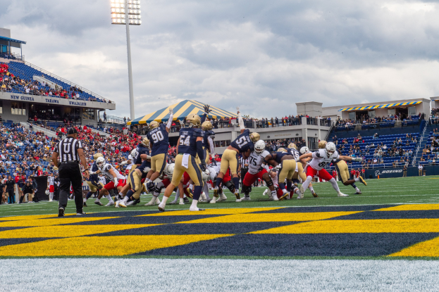 Fußballspieler in Sportkleidung und Helmen konkurrieren auf einem Feld mit Zuschauern, Stadionstrukturen und Text am Boden.