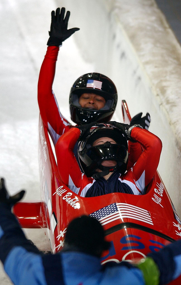 Zwei Bobsleighs bei den Olympischen Spielen, eines mit Helm und Handschuhen, mit einer Person unten und einer Wand im Hintergrund.