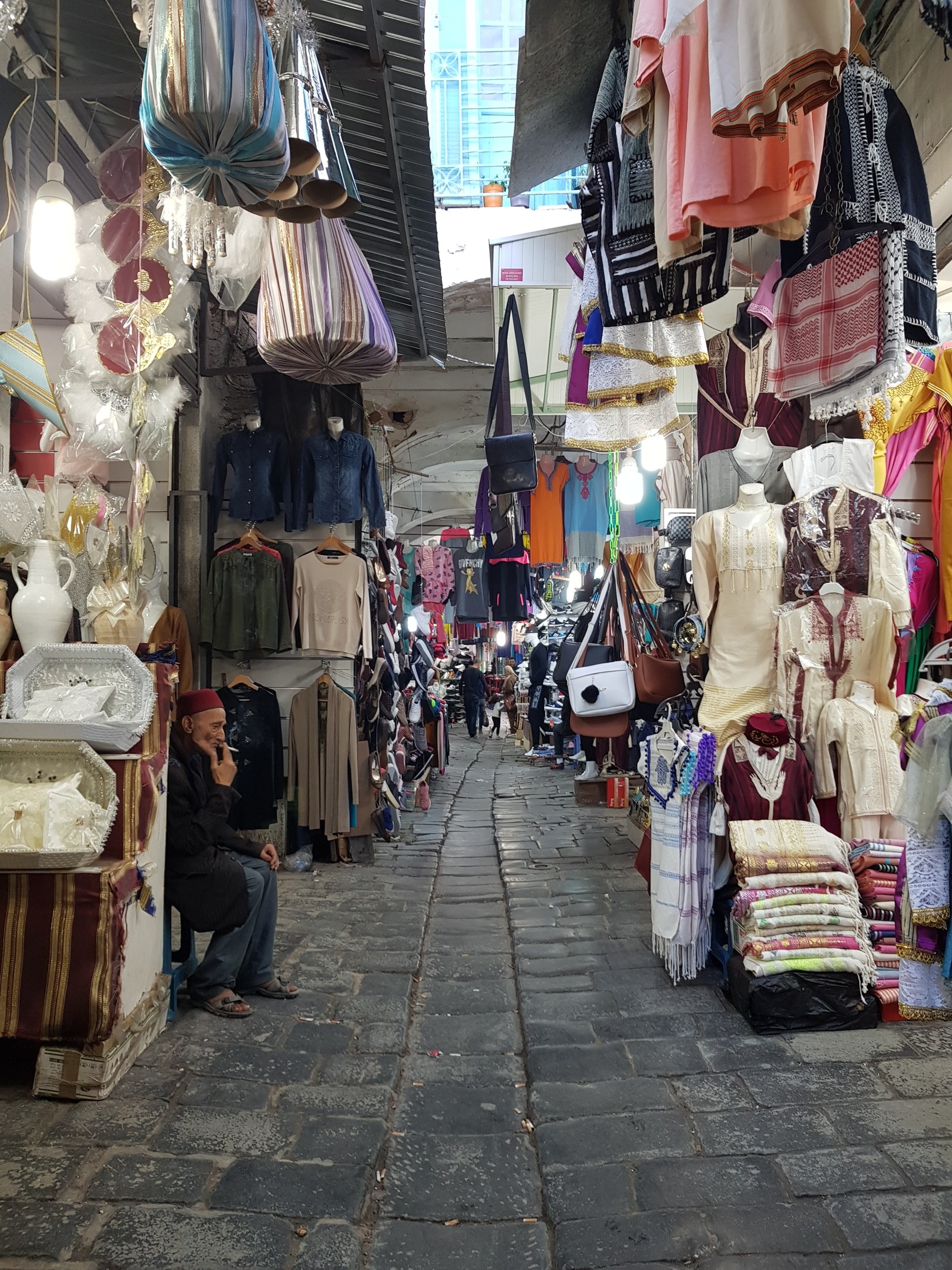 Ein belebter Straßenmarkt in der Altstadt von Jerusalem mit Ständen, die Kleidung, Taschen, Lampen und andere Artikel verkaufen, und Menschen, die die Auswahl durchstöbern.