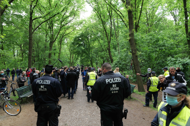 Eine Gruppe von Polizisten steht vor einer Menge während einer Anti-Terror-Demonstration in Berlin, mit Fahrrädern und einer Bank im Vordergrund und Bäumen und Himmel im Hintergrund.