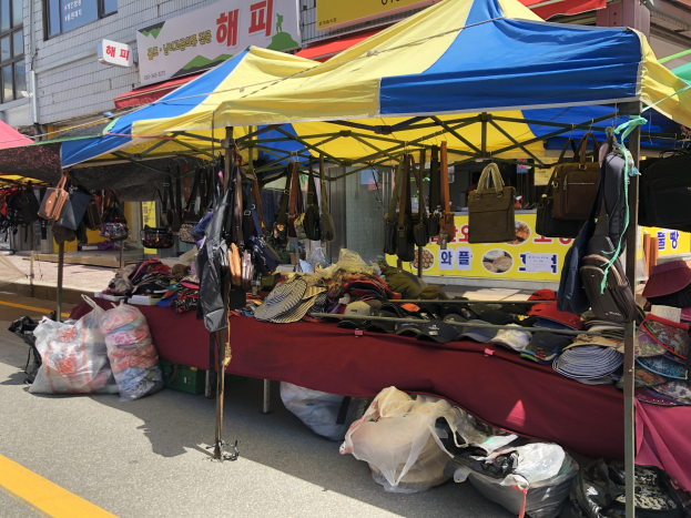 Ein Straßenmarkt mit Zelten, die Taschen, Hüte und andere Artikel präsentieren, neben Gebäuden mit Fenstern und Texttafeln.