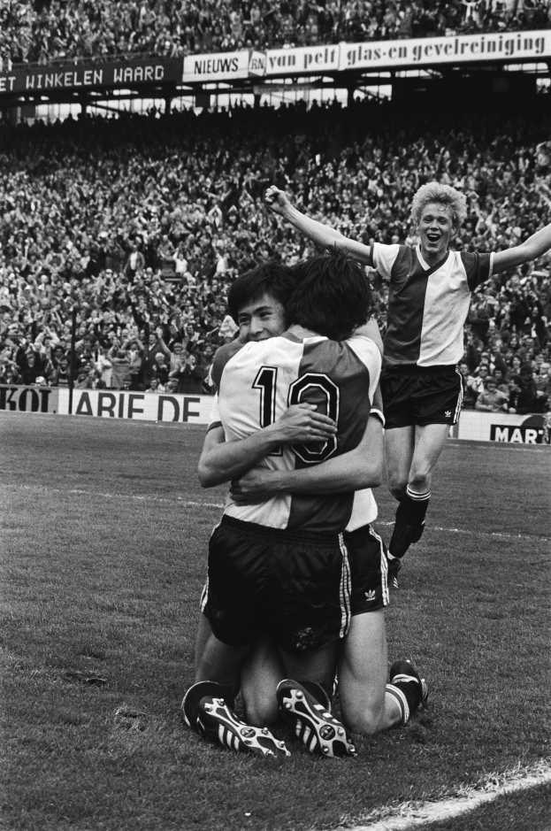 Drei Fußballspieler umarmen sich auf dem Boden nach einem Torerfolg, mit jubelnder Menge und schwenkenden Fahnen in einer schwarz-weißen Stadionatmosphäre.