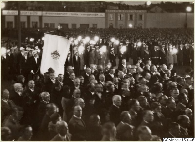 Schwarzes und weißes Foto einer großen Menschenmenge vor einem Gebäude, mit einer Person, die eine Fahne hält, Pfosten und Lichter im Hintergrund und dem Text "Gustav Klimts Rede an der Berliner Mauer" unten.