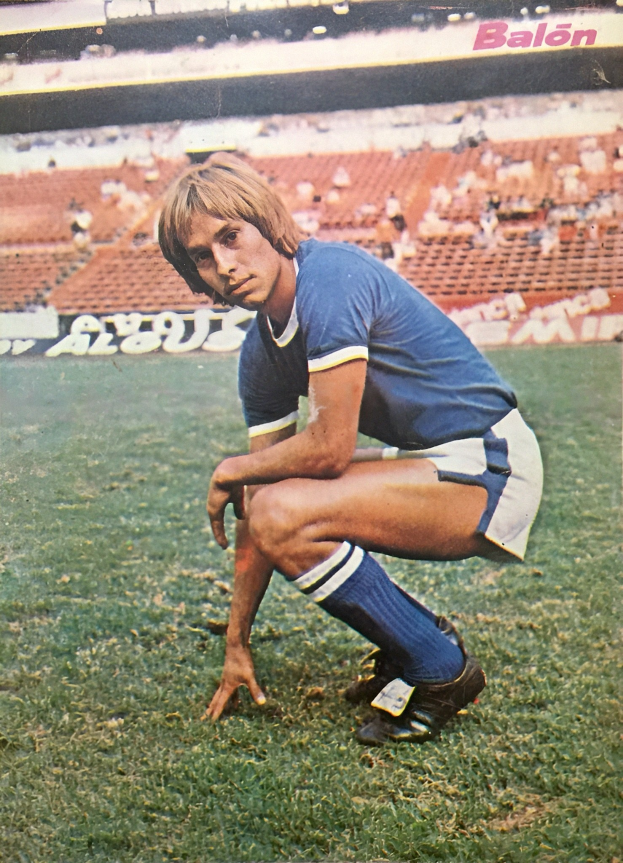 Ein Mann kniend auf einem Fußballfeld umgeben von Menschen auf Stühlen und Tafeln mit Text, trägt Schuhe.