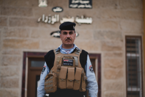 Ein Polizeibeamter in Uniform und mit Mütze steht vor einem Gebäude mit Türen, Fenstern und Text an der Wand.