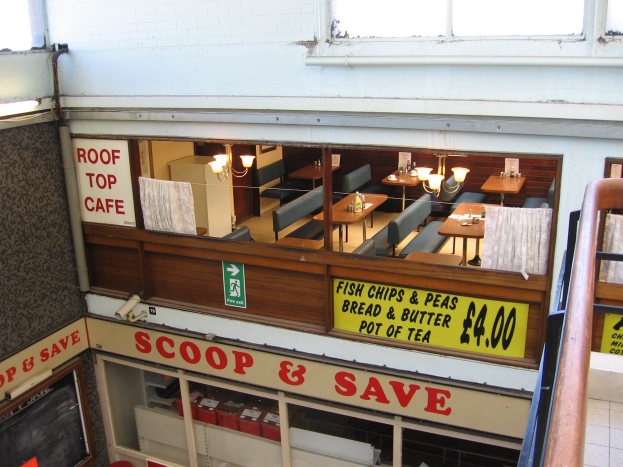 Ein Café-artiges Restaurant mit Tischen, Stühlen und beleuchteten Tafeln mit Text, das eine Brüstung auf der rechten Seite und eine Wand im Hintergrund mit der Aufschrift "Scoop & Save" hat.