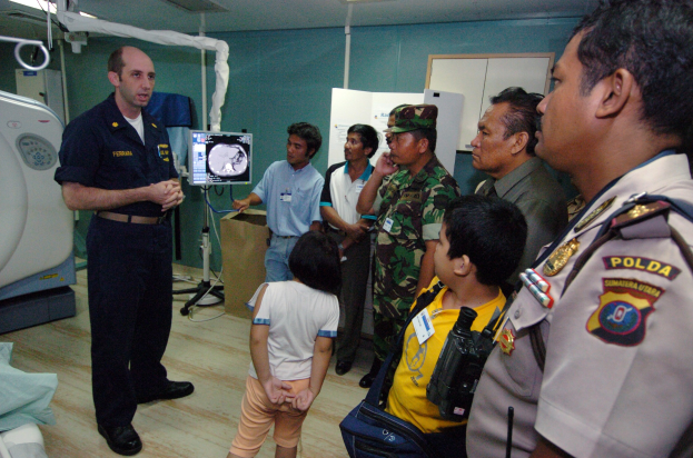 Gruppe von Menschen in einem Krankenhauszimmer mit einem Mann, der in ein Mikrofon spricht, einem drapierten Bett auf der linken Seite, medizinischer Ausrüstung im Hintergrund, einem Röntgenmaschinenständer, einer Texttafel, einer Tür und Deckenleuchten.