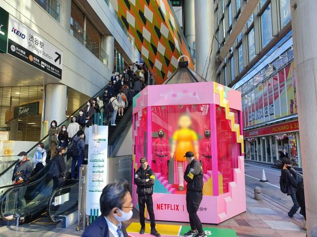 Eine Gruppe von Menschen steht vor einem modernen Gebäude mit Glaswänden und Texttafeln, einige tragen Masken, mit einer Rolltreppe im Blick; eine rosafarbene Box in der Mitte zeigt Schaufensterpuppen und trägt die Aufschrift "Netflix's new Netflix Booth in Tokyo."