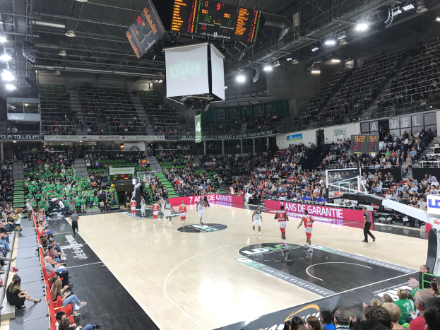 Ein Basketballspiel in einer großen Arena mit Zuschauern auf den Rängen, Anzeigetafeln mit 'Asvel vs Galatasaray' und einer Decke mit Lampen und einem Bildschirm.