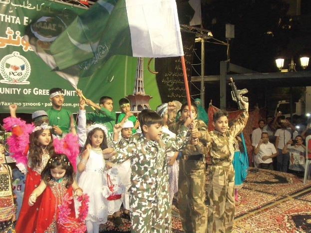 Eine Gruppe von Kindern, die Flaggen auf einem Teppichboden mit einem Banner, Lichtern und Menschen mit Kameras im Hintergrund halten, was eine Pakistan-Tag-Feier vermuten lässt.