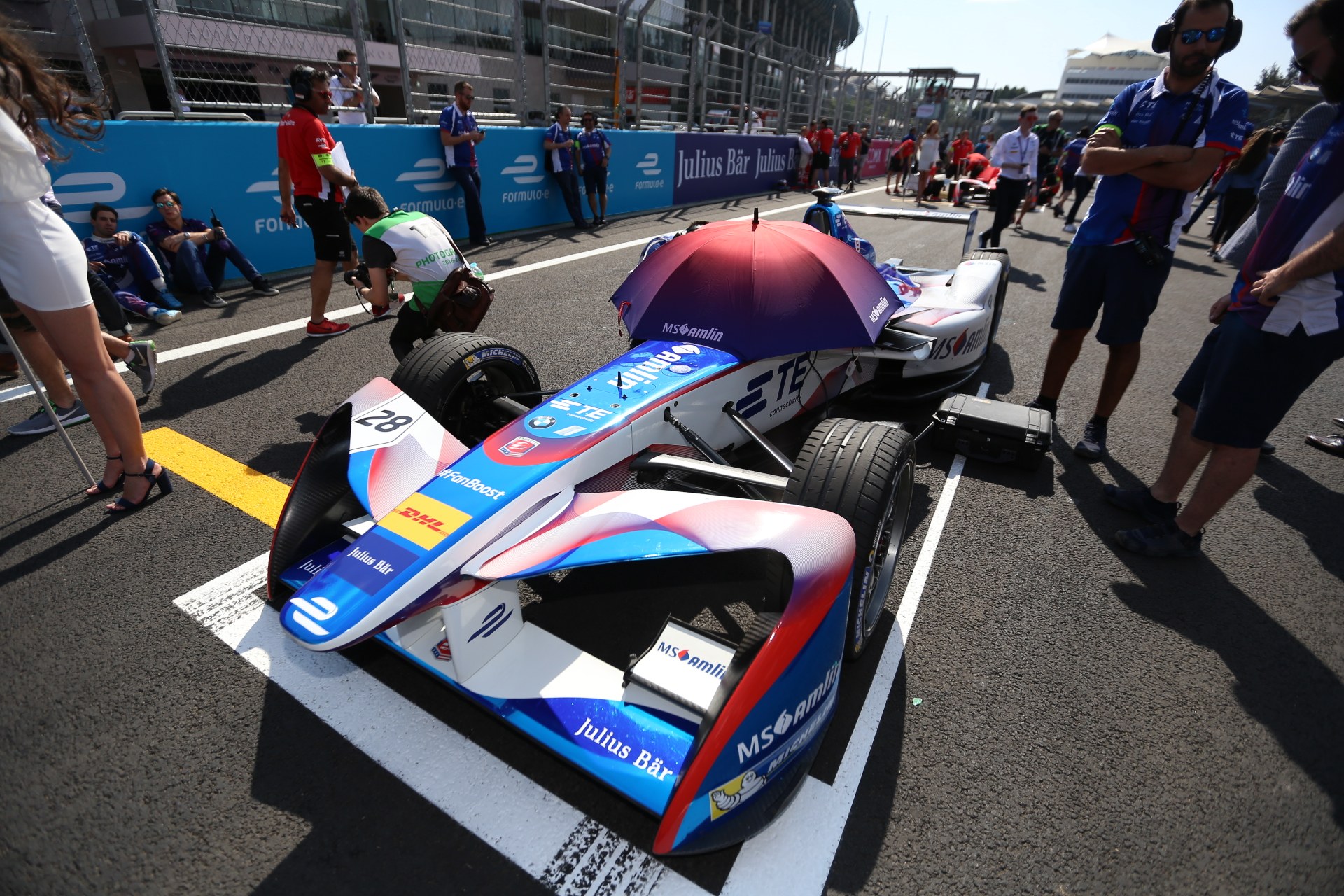 Formula E-Rennwagen mit weißer, blauer, roter und schwarzer Lackierung auf einer Rennstrecke geparkt, umgeben von Menschen, mit einem Schirm im Vordergrund und Renninfrastruktur im Hintergrund unter einem klaren Himmel.