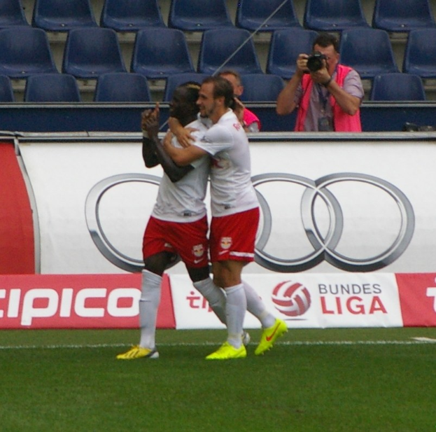 Zwei Fussballspieler feiern ein Tor auf einem Feld mit Zuschauern, einem Fotografen, einer Fahne, Stühlen und einer Wand im Hintergrund.