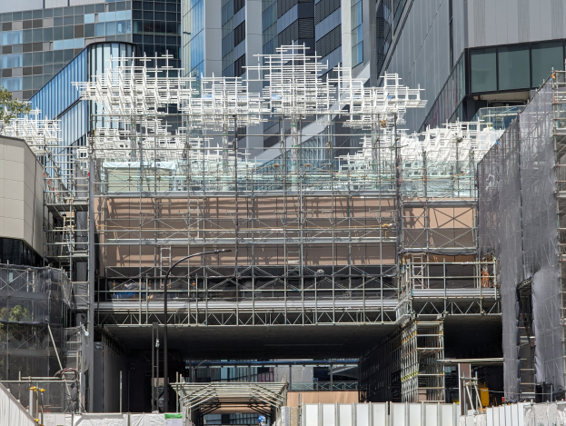 Gro├če Baustelle mit Gerüst, umgeben von B├Ąumen links und einer Stra├če unten in einer urbanen Umgebung.