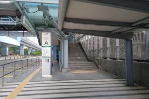 Bahnsteig mit Treppe, Straße mit Geländer auf der linken Seite, Hintergrund aus Bäumen, Pfählen, Gebäuden und einer Brücke sowie Bahnsteigdetails wie Rohre und ein Schild.