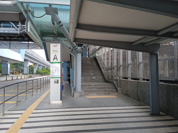 Bahnsteig mit Treppe, Straße mit Geländer auf der linken Seite, Hintergrund aus Bäumen, Pfählen, Gebäuden und einer Brücke sowie Bahnsteigdetails wie Rohre und ein Schild.